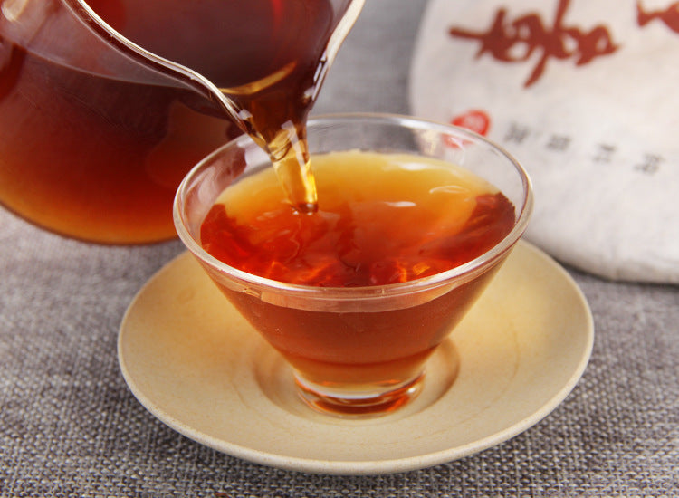 Aged ripe Pu-erh tea pouring into a tasting cup beside traditional wrapper