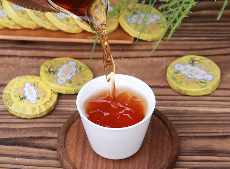 Amber-red chrysanthemum ripe Pu-erh tea liquor being poured beside wrapped mini tea cakes