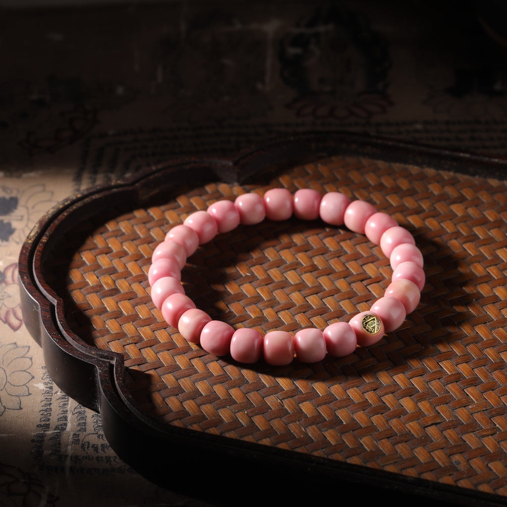 Pink Longji Bodhi barrel bead bracelet on woven tea tray with a small gold-tone marker bead