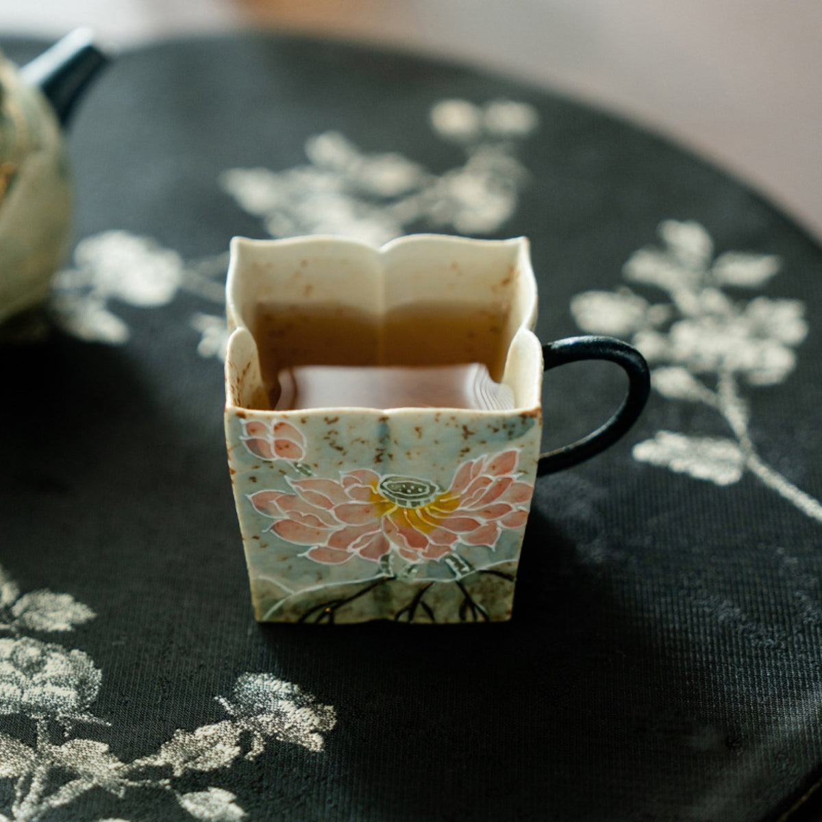 Square ceramic tea cup with pink lotus motif filled with tea on a dark tea mat