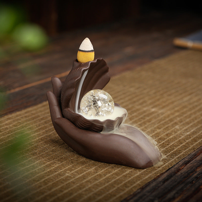Open palm crystal-orb style purple clay backflow incense burner in a tea-room setting