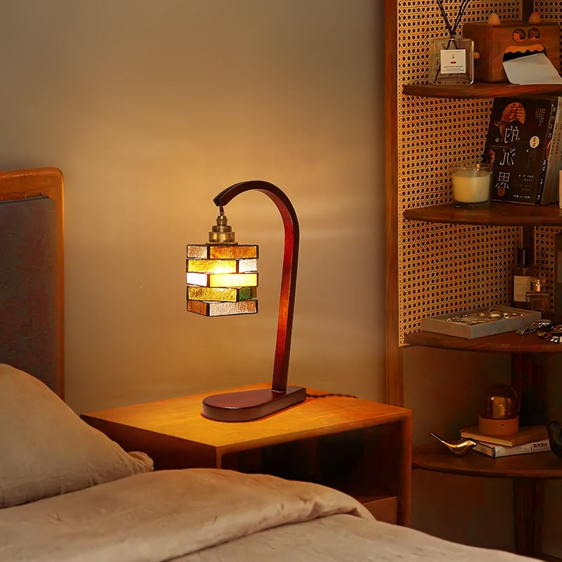 Warm stacked colored glass table lamp with curved walnut-finish arm beside a bed